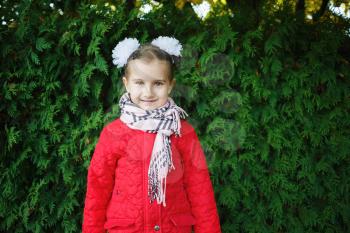 Smiling baby girl in a red jacket with white bows on her head. Child posing on a background of green arborvitae shrubs.