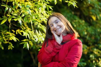 Portrait of pretty young smiling woman in a red jacket on a background of green foliage. Selective focus.