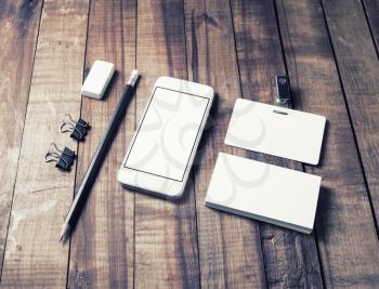 Photo of smartphone with blank white screen, business cards, badge, pencil, eraser and metal clamps on wooden table background. Blank stationery set.