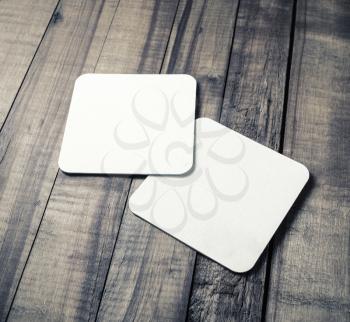 Two blank white square beer coasters on wood table background.
