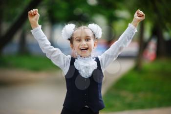 Happy child girl in school uniform with bows on head outdoors. Schoolgirl smiling and raise her hands.