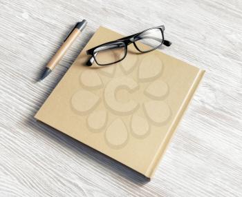 Closed blank square notebook, glasses and pen on light wood table background.