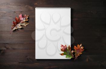 Blank spiral sketchbook and autumn leaves on dark wooden background. Mockup for your design. Flat lay.
