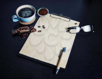 Stationery set and coffee. Clipboard with blank kraft paper, coffee cup, coffee beans, pen, glasses and ground powder on black table background.