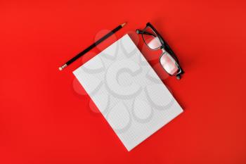 Stationery mock up. Blank copybook, glasses and pencil on red paper background. Flat lay.