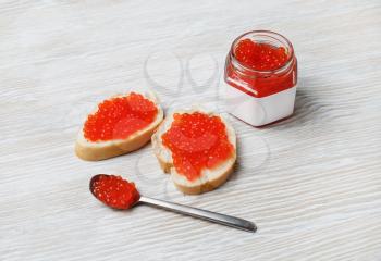 Still life with red caviar. Sandwiches with red caviar, glass jar and spoon on light wood table background.