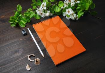 Photo of blank stationery template. Orange sketchbook, pencil, sharpener and spring flowers on wood table background.
