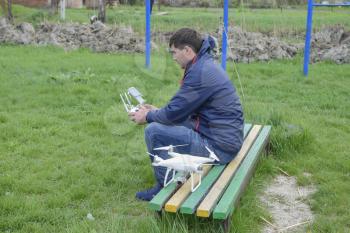 A man with a remote control quadroopter in his hands is sitting on a bench. White quadroopter prepare for flight