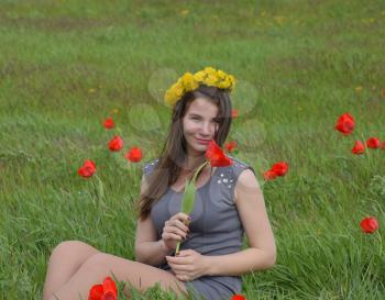 A girl with a wreath of dandelions on her head. Beautiful fairy young girl in a field among the flowers of tulips. Portrait of a girl on a background of red flowers and a green field. Field of tulips.
