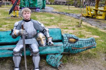 Knight in armor among wooden idols on a wooden bench. Knightly armor and weapon. Semi - antique photo.