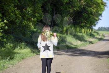 A girl walking along a forest road with a bouquet of chamomiles.