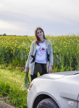 A beautiful young woman with a bouquet of daisies stands near a silver car.