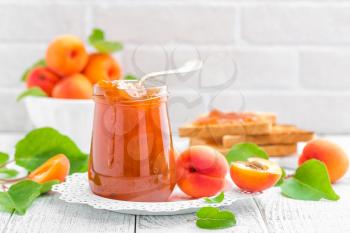 Apricot jam in a jar and fresh fruits with leaves on white wooden table, breakfast