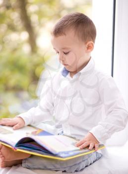 Little boy is reading book while sitting on windowsill, indoor shoot