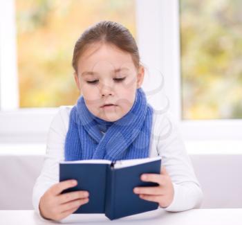 Cute little girl is reading book while sitting at table, indoor shoot