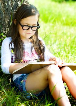 Cute little girl is reading a book while sitting on green grass