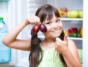 Happy girl eating plums standing near refrigerator with fruits and vegetables