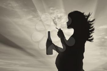 Concept of pregnancy and bad habits. Silhouette of a pregnant woman smokes a cigarette with an alcoholic drink at sunset