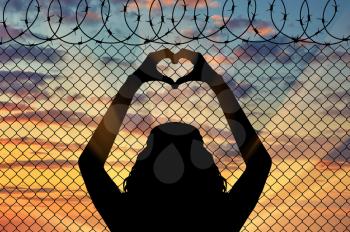 Concept of the refugees. Silhouette of a refugee shows a gesture from the hands of the heart on the background of the fence near the border