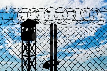 Silhouette of a watchtower and an iron fence with barbed wire on the cloudy sky