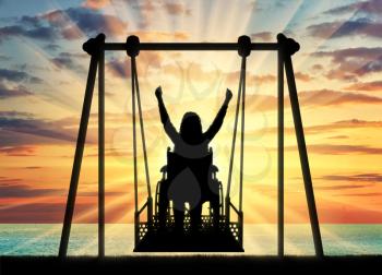 Silhouette of a happy woman is a disabled person in a wheelchair on an adaptive swing against the backdrop of a sea sunset. Concept of the lifestyle of people with disabilities