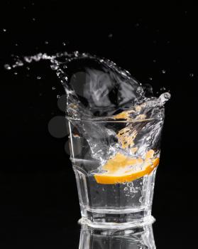 Slice of lemon splashing into a glass of water with a spray of water droplets in motion suspended in the air above the glass on a dark background.