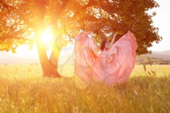 Young woman standing on a wheat field with sunrise on the background. Beauty Romantic Girl Outdoors. The dress fluttering in the wind at sunset. flying fabric.