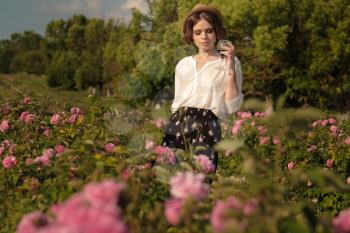 Beautiful young woman with curly hair posing near roses in a garden. Girl with a glass in her hand, tasting red wine and having fun