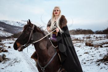 Beautiful young blonde on a crow. Woman viking with a black horse against the background of mountains