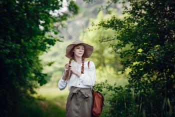 Lovely young woman with red hair, in a hat and with a backpack on the edge of the forest. Beautiful red-haired girl on a walk