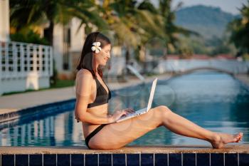 Woman using her laptop on the pool edge on a sunny day. Freelance work in tropical country