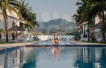 Woman doing yoga exercise at poolside. Young attractive woman practicing yoga in lotus pose. Healthy, beauty concept, reflection on water, meditating at sunrise.