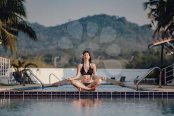 Woman doing yoga exercise at poolside. Young attractive woman practicing yoga in lotus pose. Healthy, beauty concept, reflection on water, meditating at sunrise.