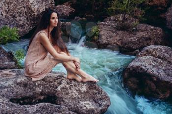 Young woman in the dress is sitting on the stone in the middle of a creek. Fashionable toning.