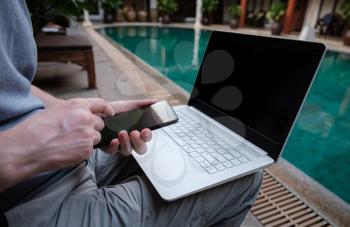 travel blogger sitting at swiming pool with white laptop and smartphone, online banking on internet in travel. Freelance remote work concept. Copy space, sea view background