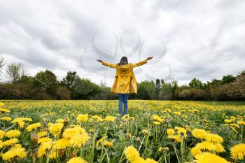 Portrait of a beautiful woman in a yellow cloak with arms raised on a field of flowering dandelions. Stormy sky in spring