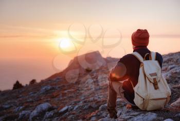 A young man sitting on top cliff in spring mountains at sunset and enjoying view of nature. Mountain and coastal travel, freedom and active lifestyle