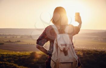 Pretty traveling woman standing on top of mountain at sunset and using mobile phone. High mountains touristic path. Digital communication.