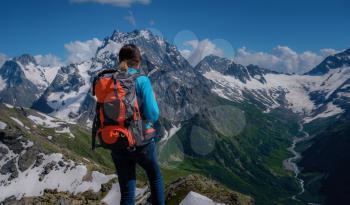 A young woman tourist in a blue blouse with an orange hiking backpack enjoys a breathtaking view of the mountains. North Caucasus, Dombai, Russia. mountaineering sport lifestyle concept