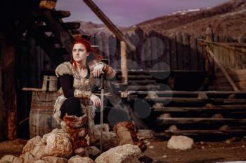 Viking woman with red hair. In the hands of the sword to fight the enemy. Reconstruction of a medieval scene. Against the backdrop of a large viking village.