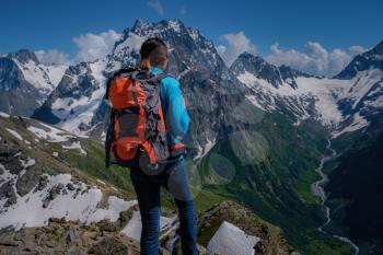 A young woman tourist in a blue blouse with an orange hiking backpack enjoys a breathtaking view of the mountains. North Caucasus, Dombai, Russia. mountaineering sport lifestyle concept