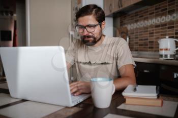 Focused young man wearing glasses using laptop, typing on keyboard, writing email or message, chatting, shopping, successful freelancer working online on computer, sitting in kitchen