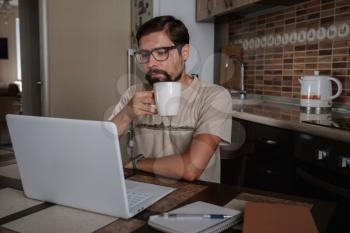 Young entrepreneur sitting at table drink coffee work at laptop thinking of problem solution, thoughtful male employee pondering considering idea looking at computer screen making decision