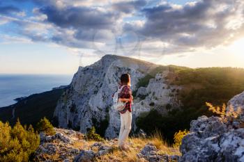 Fit female hiker with backpack standing on a rocky mountain ridge looking out of green alpine valleys and peaks in a healthy outdoors lifestyle concept