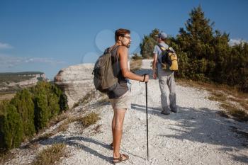 Two Men walking on rocky slope carrying Backpacks using trekking Sticks. Mountains valley View beside of People. summer travel to nature. Hiker in background.