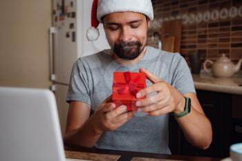 A hipster man with a red cup sitting on a sofa at home at Christmas time. Virtual Christmas house party. Online team meeting video conference calling from home. Man wearing Santa hat