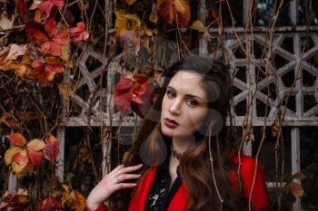 Beautiful young woman in red coat is enjoying autumn park. A pleasant walk on the background of a fence with branches of wild grapes and red leaves