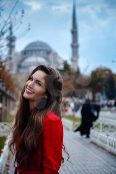 Latin American woman or Turkish woman in a red stylish coat in front of the famous blue mosque in Istanbul, Photo of traveler on the background of a mosque on autumn day.
