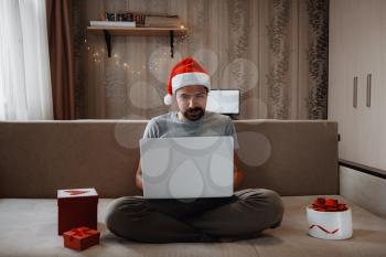 A hipster man with a red cup sitting on a sofa at home at Christmas time. Virtual Christmas house party. Online team meeting video conference calling from home.