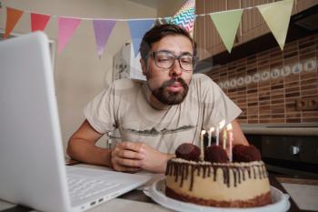 Man celebrating birthday online in quarantine time. Guy celebrating his birthday , blowing out the candle on the birthday cake and making video call. Coronavirus outbreak 2020.